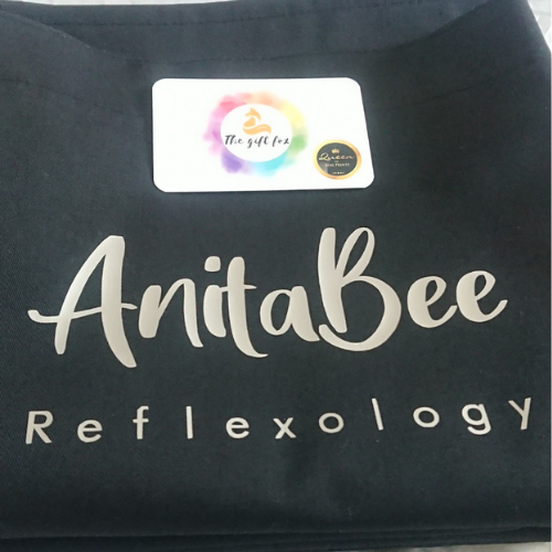 Black apron folded with anita Bee reflexology printed on the front in silver. 